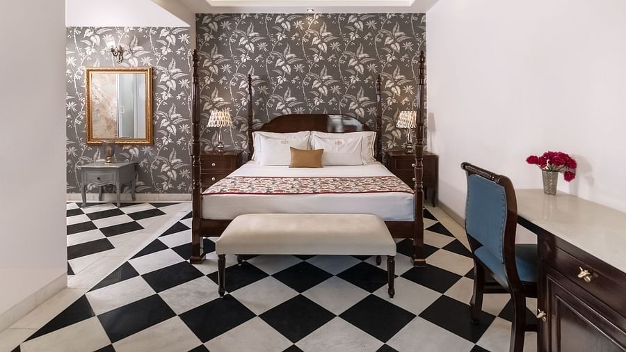 A bedroom with a four-poster bed, a patterned wallpapered wall, a chequered black and white tiled floor, and a writing desk with a blue chair | Dileep  Kothi, Jaipur